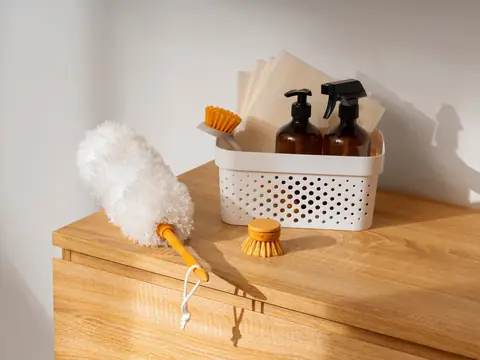 A white basket holding brown pump and spray bottles, folded cloths and a scrubbing brush on a wooden cabinet, with a fluffy white duster beside it