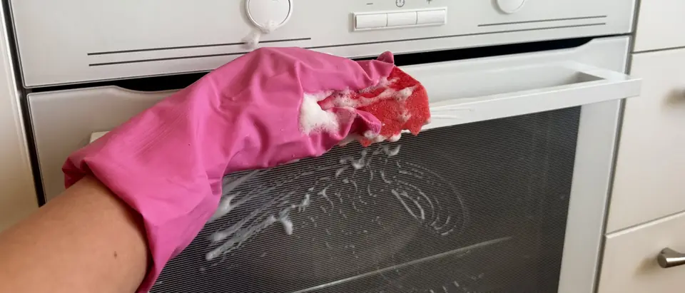A hand in a pink rubber glove scrubbing the glass door of a white oven with a soapy red sponge