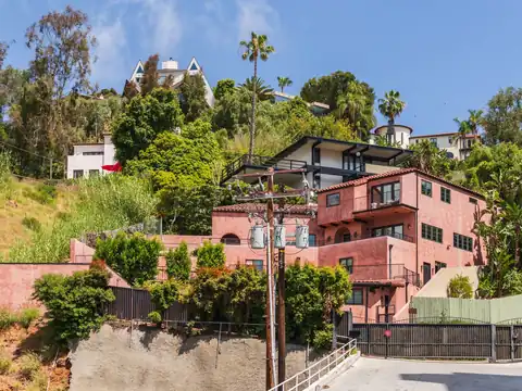 A pink stucco house with a red tile roof and other houses stepping up a steep green hillside with palm trees, above a concrete road and a gated driveway