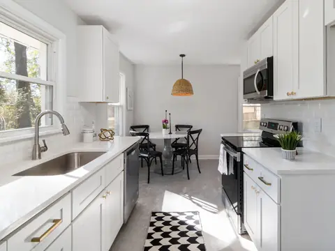 A bright white kitchen with a stainless steel sink, clear stone counters and an electric range, and a small dining table at the far end