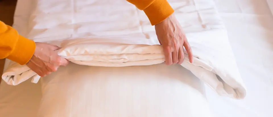Two hands in orange sleeves folding a white duvet back over a pillow on a freshly made bed