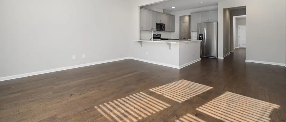 An empty apartment living room with dark wood floors and an open kitchen, with sunlight falling through blinds onto the floor