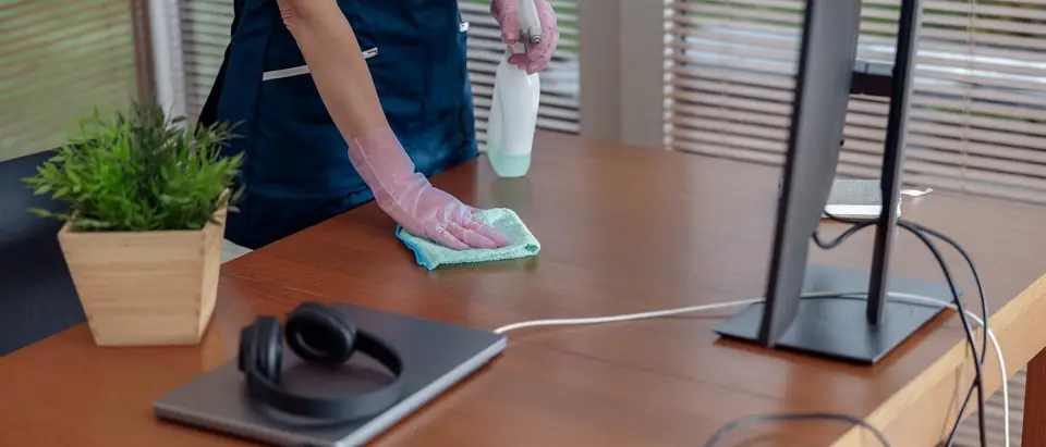 A gloved hand wiping a wooden office desk with a green cloth beside a spray bottle, with a monitor and a laptop on the desk