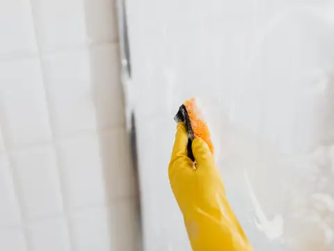 A hand in a yellow rubber glove washing a soapy glass shower panel with an orange sponge, beside white wall tiles